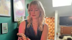 Jess Davies sentada em casa olhando para o celular. Pode-se ver uma TV sobre um móvel atrás dela e uma parede azul-turquesa com um quadro emoldurado à sua esquerda.