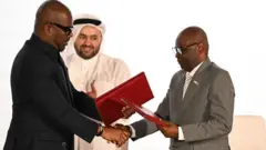 Sumbu Sita Mambu, intumwa nkuru ya DRC mu biganiro by'i Doha, asuhuza n'umunyamabanga nshingwabikorwa wa M23 Benjamin Mbonimpa, muri Hoteli Sheraton, i Doha muri Qatar, nyuma yo gusinya ku nyandiko y'imiterere-remezo y'amasezerano y'amahoro. Sumbu yambaye ikositimu y'umukara, naho Mbonimpa yambaye ikositimu ya khaki. Iruhande rwabo inyuma hari intumwa ya Qatar irimo gukoma amashyi, imwenyura. Ifoto yo ku itariki ya 15 Ugushyingo (11) mu mwaka wa 2025. 