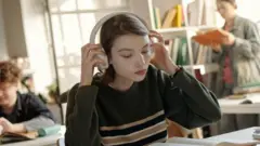 Młoda kobieta zakłada słuchawki, czytając książkę i przygotowując się do egzaminów w bibliotece szkolnej; wolne miejsce na tekst.
