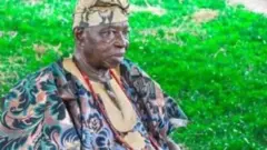 Oloye Lere Paimo jókòó sórí àga nínú aṣọ Damaski agbada àti fìlà to jẹ́ aláwọ̀ goolu àti omi aro. Ó sì tún fi ìlẹ̀kẹ̀ si ọrùn nígbà tí àwọn koríko rẹsurẹsu wá ní ilẹ ibi tí wón jókòó sí. 