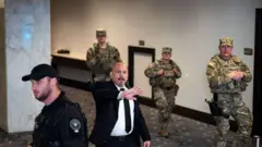 ホテルの廊下を迷彩服を着た武装警備員3人が歩いている。その横で、黒いスーツを着てイヤホンを耳に付けた人が、カメラに向かって後退するよう右手を広げている