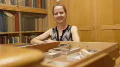 A paleontóloga Emma Nicholls olhando na direção da câmera, com os fósseis na frente, organizados numa mesa