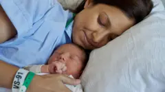 
Happy mother lying on couch with newborn baby - stock photo Smiling young woman in blue robe lying on couch with adorable newborn baby sucking hand while resting after delivery in hospital room