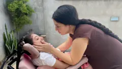 Uma mulher olhando para a filha que está em uma cadeira de rodas. Ela segura seu rosto.