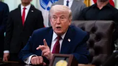 特朗普坐在白宮橢圓形辦公室的辦公桌前對記者講話，他舉起左手食指說明觀點（18/4/2026）