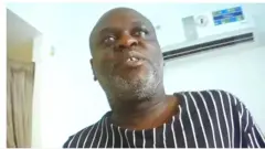 Aworan yii ṣafihan Charles Mwegiswa, eyin rẹ meji han nita, o n wo ọọkan, o wọṣo dudu to ni ila funfun tẹẹrẹtẹ