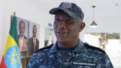 A head and shoulders image of a man in a blue camouflage uniform and peaked cap. A badge says "Eth Federal Police" on his chest and an Ethiopian flag is in the background.