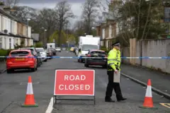 Una calle en Glasgow, Escocia, acordonada con cinta azul y blanca de la policía. Un oficial con la distintiva chaqueta fluorescente habla con alguien que está fuera del encuadre.