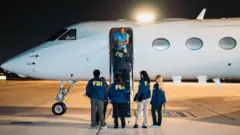 Mavi formalı bir adam elleri kelepçeli bir jetten iniyor, 4 FBI memuru arkaları dönük şekilde yerde onu karşılıyor. 