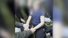 Скриншот з відео поліції, обличчя заблюрені, юнака у футболці тримає за руки інша людина, довкола стоять люди