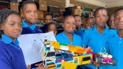 Abana benshi bahagaze bambaye imyambaro y'ishuri y'ubururu. Bafite za robo z'amabara y'umuhondo, icyatsi kibisi, umweru, umutuku n'iroza. Barareba muri 'camera', umwe muri bo arimo kumwenyura. Inyuma yabo mu ishuri hari ibikarito n'ibindi bikoresho. 