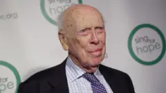James Watson em 2016, com 89 anos. Ele veste um terno escuro, camisa branca xadrez e gravata roxa, com detalhes em tom rosa e azul claro.