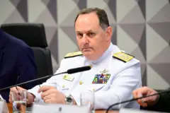 Almirante está fardado de branco. Ele olha para o lado. Na frente dele há um microfone e um copo de água em cima da mesa. Ele está sentado.