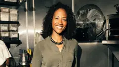 Uma mulher de cabelos cacheados pretos sorri para a câmera. Ela está em uma cozinha, ao lado de um grande ventilador. Ao fundo, há utensílios de cozinha e o cotovelo de um homem que parece estar trabalhando atrás dela,