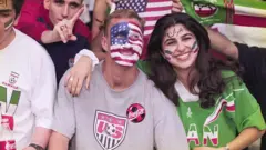 Болельщик США и болельщица Ирана стоят в обнимку, на их лицах нарсиованы национальные флаги