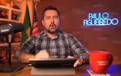 Paulo Figueiredo está em uma mesa com um notebook, um microfone e uma xícara. Atrás há uma bandeira do Brasil e dos EUA e uma logo iluminada com o nome dele. 
