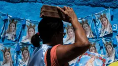 Uma mulher carrega um tijolo em cima da cabeça diante de um muro azul coberto por estampas repetidas de uma figura religiosa.