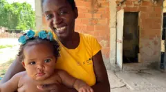 Evelyn sorrindo em pátio de casa, com bebê no colo