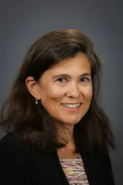 A imagem mostra uma mulher de cabelos castanhos na altura dos ombros, sorrindo para a câmera em um retrato de estúdio. Ela veste um blazer preto sobre uma blusa estampada e usa brincos pequenos, tendo ao fundo um cenário cinza neutro.