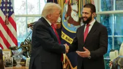 ホワイトハウスの大統領執務室の窓際でにこやかに握手するトランプ氏とアル・シャラア氏。共にダークスーツに赤いネクタイを着けている。アル・シャラア氏は長いひげを蓄えている