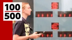 На заднем плане табло курса евро и доллара без значений. На переднем плане слева направо идет девушка со светлыми волосами, в футболке с неразборчивой надписью. В руке девушка держит телефон.