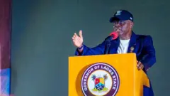 Gomina ipinlẹ Eko, Babajide Sanwo-Olu n sọrọ sinu ẹrọ gbohungbohun. O wọsọ alawọ aro, o de fila o si lo igo soju