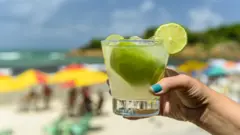 Mão segurando copo de caipirinha na praia
