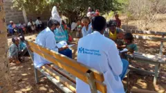 የኢንተርናሽናል ሜዲካል ኮርን አርማ የያዘ ነጭ ጋውን የለበሱ የጤና ባለሙያዎች ነዋሪዎችን መረጃ እየመዘገቡ