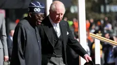 King Charles III wit di President of Nigeria Bola Ahmed Tinubu on di Royal Dais during a welcome ceremony for Datchet Road for Windsor. Both men wear black, wit King Charles wey wear three-piece suit
