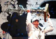Larry Ellison, vestido con gorra y ropa deportiva blanca, levanta un gran trofeo plateado con ambas manos.