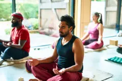 Foto colorida mostra três pessoas sentadas no chão com as pernas cruzadas e olhos fechados,times para apostarposiçãotimes para apostarmeditação