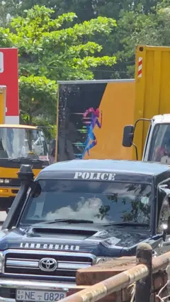 පොලිස් ජීප් රථයක් ඉදිරියෙන් ගමන් කරන අතර කහ පැහැති ලොරි තුනක්  පිටුපසින් පෙනේ.