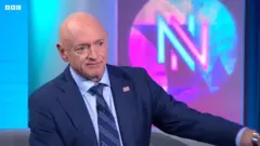 水色と赤紫のライトで「NN」と背景に描かれたBBCスタジオで座って話す、スーツ姿のケリー上院議員。襟もとに星条旗のピンバッジをつけている