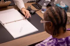 Yvonne Gregwor sentada e olhando para um papel com ilustrações sobre a mesa