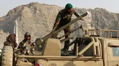 Yeşil kamuflajlı üniformalar giyen Taliban askerleri, bir askeri araca roketatar yüklüyor. Arka planda dağlar görünüyor.