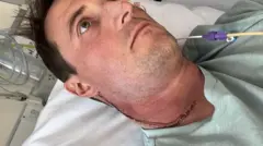 Anthony Perriam está sentado, olhando para o lado, sem encarar a câmera. Ele tem uma grande cicatriz de uma operação que percorre desde a orelha até a parte inferior do pescoço