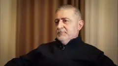 يجلس رجل على كرسي مرتدياً سترة سوداء وينظر إلى جانب الكاميرا