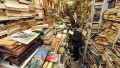 Jovem parado no meio de uma livraria antiga cercado por livros usados. 