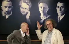 Dois homens estão em frente a um quadro de fotos que mostra 4 pessoas da família Curie