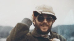 Kenner Elias Jones is a white man in his 30s or 40s in this picture, with a brown beard and moustache. He wears dark lensed prescription glasses and a cream coloured hat which he is holding, a checked shirt and tie covered by a brown coat with a collar. He is smiling at the camera. The image is grainy.