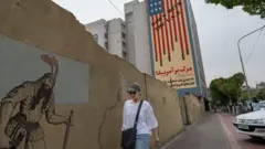 Una joven iraní camina por una acera bajo un mural antiestadounidense en el centro de Teherán, Irán.