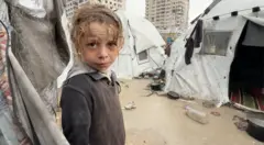 طفل فلسطيني مبلل من المطر ينظر إلى الكاميرا، محاط بخيام غارقة بالمياه وأرض موحلة ومبللة.