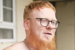 John Gladwin, de óculos e barba longa, usando um colar, com janela ao fundo, desfocada