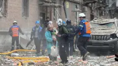 Женщина с собачкой в руках выходит из дома, на земле — обломки кирпичей, рядом с ней несколько человек в форме спасателей.