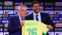 O técnico italiano Carlo Ancelotti recebe uma camisa amarela da seleção brasileira das mãos do presidente da Confederação Brasileira de Futebol (CBF), Samir Xaud, em 26 de maio de 2025, no Rio de Janeiro. Os dois aparecem sorrindo, vestidos de terno e gravata.