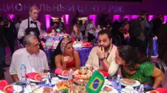 No que parece ser uma grande reunião, vemos quatro pessoas sentadas à mesa repleta de uma variedade de alimentos e bebidas. Entre os pratos, há uma pequena bandeira brasileira. Ao fundo, vemos garçons, fotógrafos, mais pessoas sentadas e conversando e uma placa com letras em alfabeto cirílico.