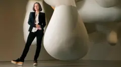 Sarah Thornton, vestida com uma blusa branca, jaqueta e calça preta, ao lado de uma obra de arte que consiste em formas gigantes de cor branca