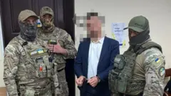 Чоловік у костюмі в кайданках із заблюреним обличчям, біля нього - троє чоловіків у камуфляжній формі із нашивками НАБУ