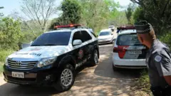 Varios vehículos policiales en la granja tras la muerte de Victoria. Un hombre uniformado observa los vehículos desde la carretera.