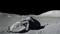Uma foto em preto e branco de uma enorme rocha na superfície lunar, que faz um astronauta caminhar ao seu lado parecer minúsculo. Ao fundo, vê-se o veículo do astronauta. Montanhas são visíveis contra um "céu" completamente negro.